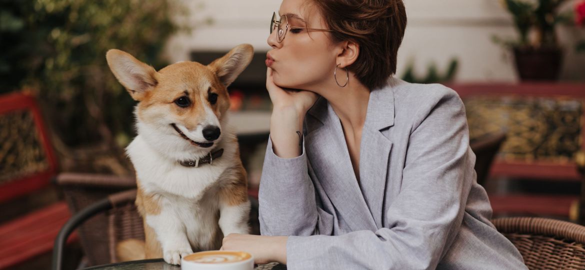 Cool girl in grey suit kisses her dog and chills in street cafe. Pretty dark-haired woman in jacket